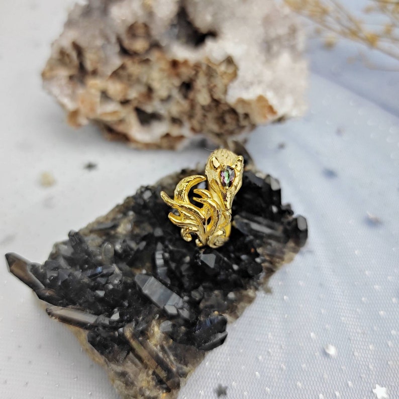 May include: A gold ring with a detailed design of a mythical creature with a green gemstone eye. The ring is displayed on a bed of black crystals.