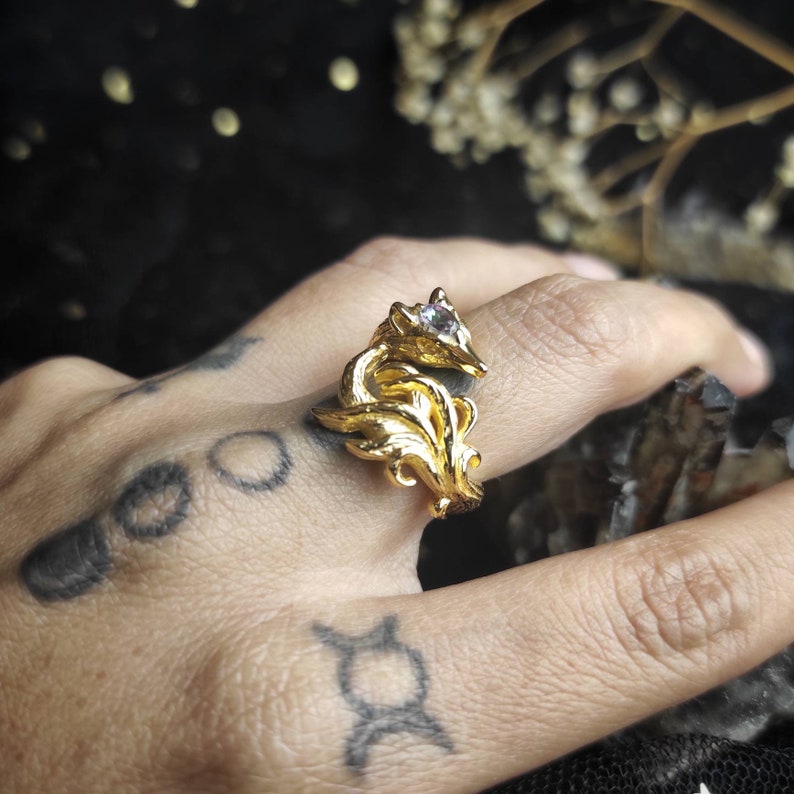 May include: A gold-toned ring featuring a detailed fox design with a clear gemstone eye. The ring is worn on a hand with tattoos.