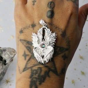 Peut inclure: Pendentif en argent avec un cœur ailé et un seul œil au centre. L'œil est une pierre précieuse irisée avec un arc-en-ciel de couleurs.
