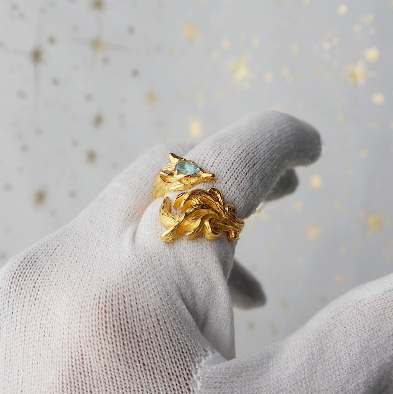 May include: A gold ring with a fox design and a blue gemstone in the eye. The ring is on a white surface.