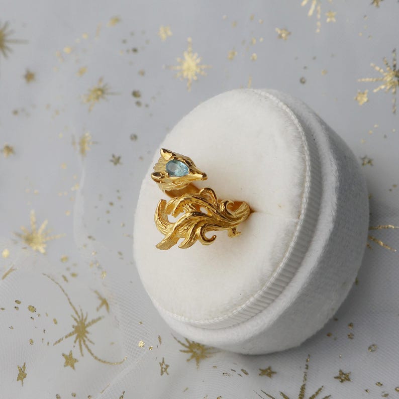 May include: A gold ring featuring a detailed fox design with a blue gemstone for an eye. The ring is displayed on a white velvet ring box.