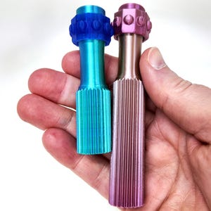 May include: Two decorative bottle stoppers. One is teal and blue, the other is pink and bronze. Both have a ribbed body and a decorative top. The stoppers are held in a hand.