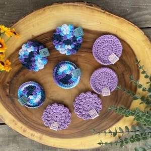 May include: Eight crocheted coasters in shades of purple and blue with the text "Handmade with Love" on each. The coasters are arranged on a wooden surface.