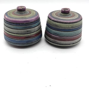 Op de afbeelding: Twee keramische potten met deksels. Elke pot is cilindrisch met een afgeronde bovenkant en een kleine knop. De potten zijn versierd met horizontale strepen in verschillende kleuren, waaronder paars, groen, blauw en rood. De potten staan op een witte achtergrond.