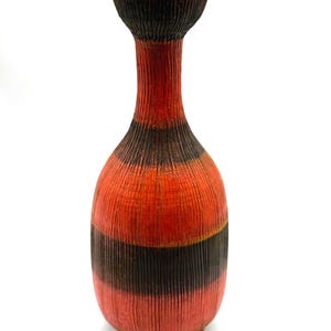 May include: A textured ceramic vase in a distinctive shape. It features bands of red and dark brown. The top has a flared opening. The vase has a ribbed texture. A decorative item for the home. The vase is approximately 25 cm tall.