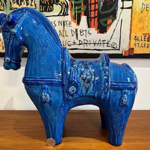 May include: A blue ceramic horse figurine with a saddle and decorative details. The horse is standing on a wooden surface.