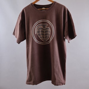 May include: A brown t-shirt with a tan grenade graphic on the front. The grenade is inside a circle with a tan outline.