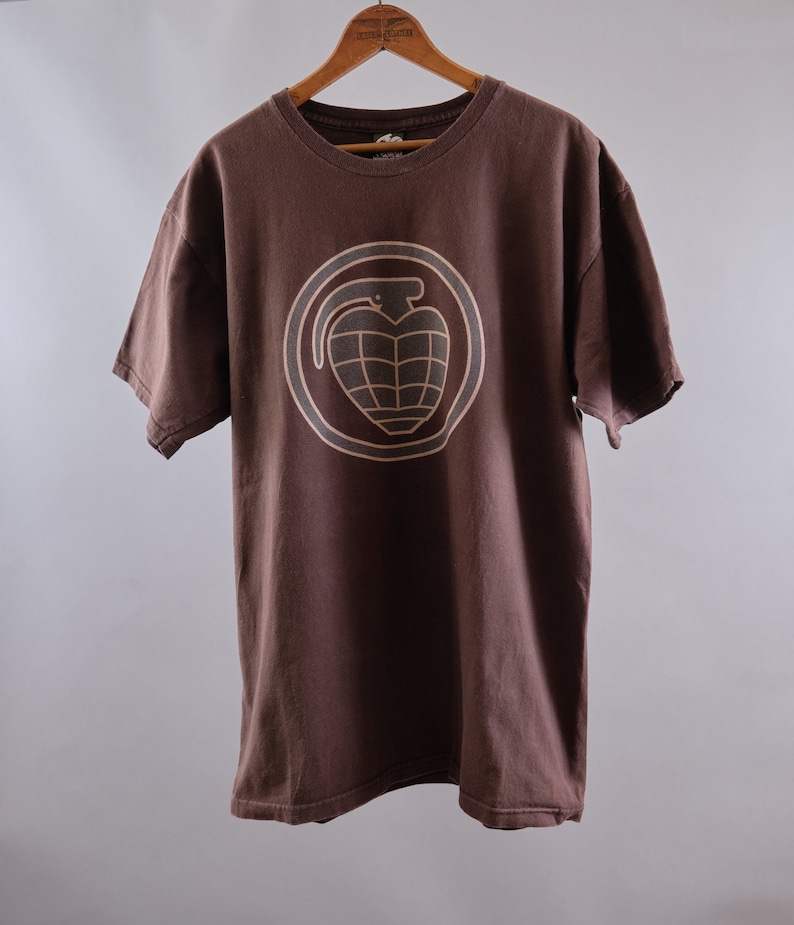 May include: A brown t-shirt with a tan grenade graphic on the front. The grenade is inside a circle with a tan outline.