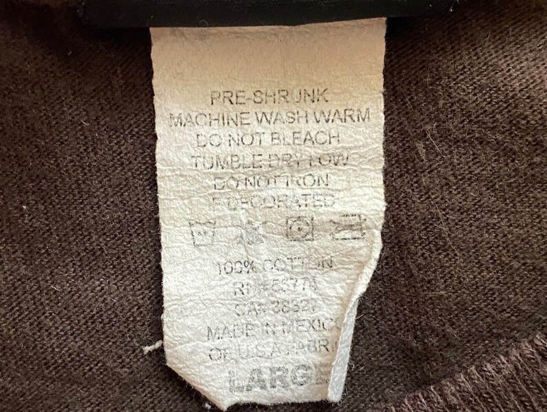 May include: A white care label with black text detailing washing instructions for a garment. The text reads "Pre-shrunk, Machine wash warm, Do not bleach, Tumble dry low, Do not iron, Decorated, 100% Cotton, RN 38327, Made in Mexico, Large."
