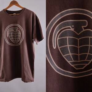 May include: Brown t-shirt with a tan graphic of a grenade in a circle. The grenade has a heart shape.