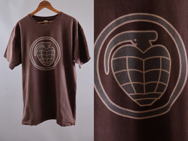 May include: Brown t-shirt with a tan graphic of a grenade in a circle. The grenade has a heart shape.