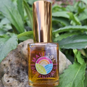 May include: A small, clear glass bottle with a gold cap. The bottle contains a golden liquid and has a label with the text "Maui Sol Botanicals" and a colorful logo of a sun, waves, and a leaf.