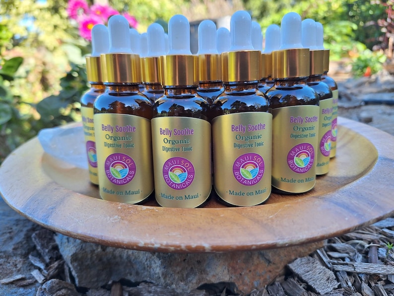 May include: A collection of small brown glass bottles with white droppers, labeled "Belly Soothe Organic Digestive Tonic." The bottles are arranged on a wooden plate, with the text "Maui Sol Botanicals" and "Made on Maui" visible.