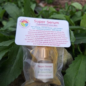 May include: A small clear bottle with a gold cap containing a serum labeled "Super Serum Age Defying Anti-Oxidant Oil Blend 100% Natural & Organic". The bottle is packaged in a clear plastic bag with a gold mesh lining. The bottle is sitting on a rock with green foliage in the background.