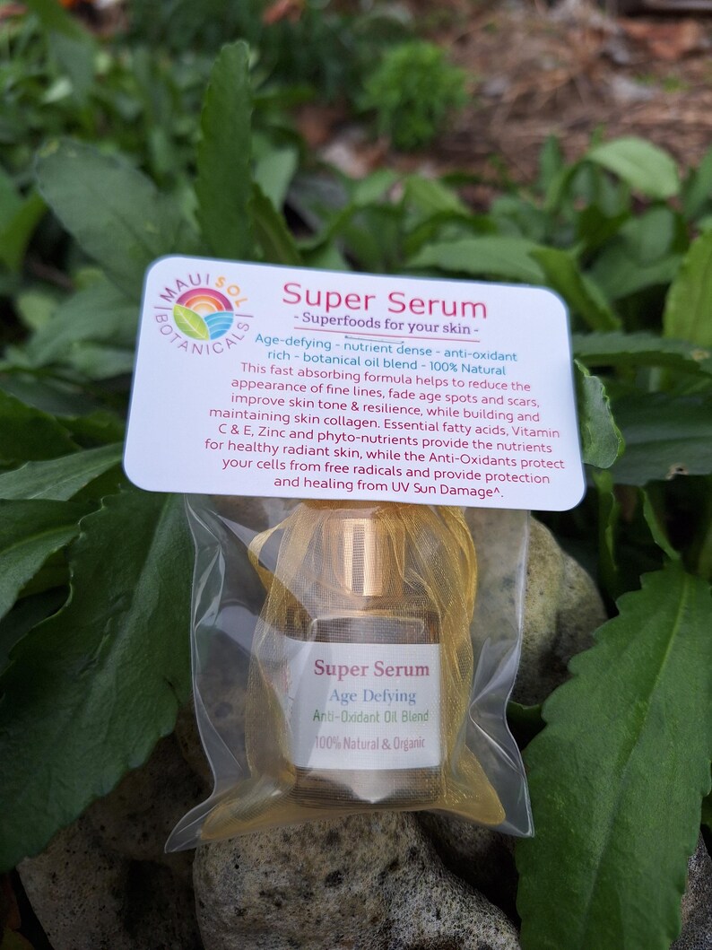 May include: A small clear bottle with a gold cap containing a serum labeled "Super Serum Age Defying Anti-Oxidant Oil Blend 100% Natural & Organic". The bottle is packaged in a clear plastic bag with a gold mesh lining. The bottle is sitting on a rock with green foliage in the background.