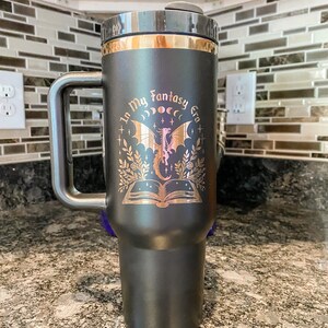 Puede incluir: Taza de viaje negra con asa y tapa doradas. La taza tiene un dise&ntilde;o dorado de un drag&oacute;n, un libro y el texto "In My Fantasy Era".