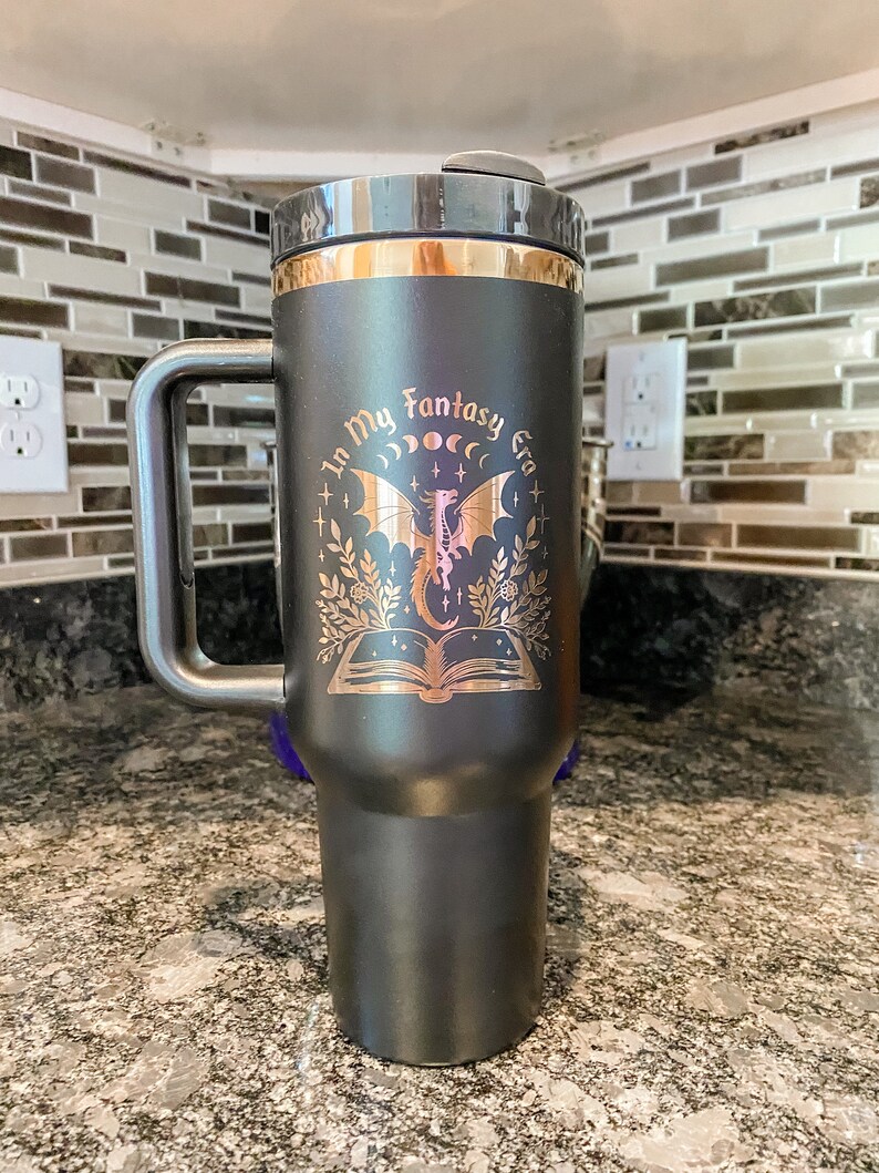 Puede incluir: Taza de viaje negra con asa y tapa doradas. La taza tiene un dise&ntilde;o dorado de un drag&oacute;n, un libro y el texto "In My Fantasy Era".