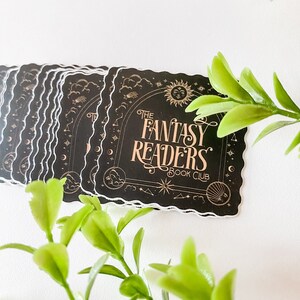 K&ouml;nnte beinhalten: Ein Stapel schwarzer und goldener Aufkleber mit einer gewellten Umrandung. Der Aufkleber zeigt einen Halbmond, Sterne und Wolken mit dem Text "The Fantasy Readers Book Club".