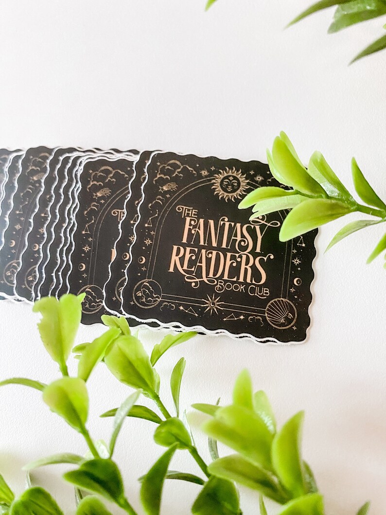 K&ouml;nnte beinhalten: Ein Stapel schwarzer und goldener Aufkleber mit einer gewellten Umrandung. Der Aufkleber zeigt einen Halbmond, Sterne und Wolken mit dem Text "The Fantasy Readers Book Club".