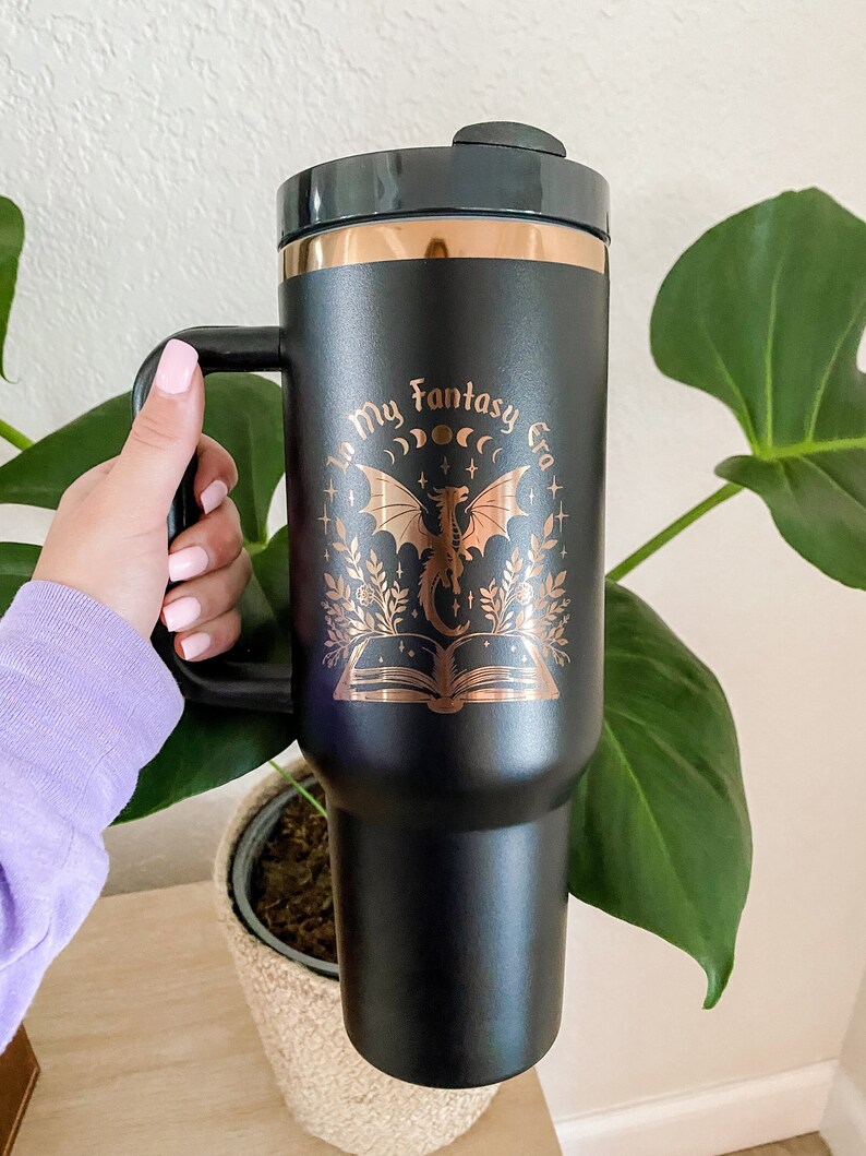 Puede incluir: Taza de viaje de acero inoxidable negro con un dise&ntilde;o dorado que presenta un drag&oacute;n, un libro y el texto "In My Fantasy Era".