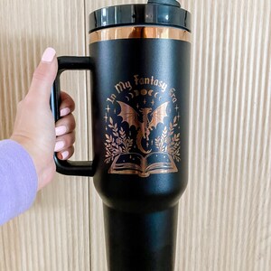 Puede incluir: Taza de viaje negra con un dise&ntilde;o de color cobre que presenta un drag&oacute;n, un libro y el texto "In My Fantasy Era".