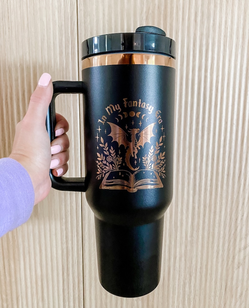 Puede incluir: Taza de viaje negra con un dise&ntilde;o de color cobre que presenta un drag&oacute;n, un libro y el texto "In My Fantasy Era".