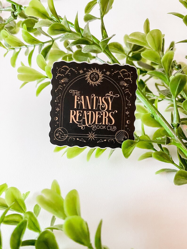 K&ouml;nnte beinhalten: Schwarzer und goldener Aufkleber mit einem dekorativen Rand. Der Aufkleber zeigt einen Halbmond, Sterne und eine Sonne mit einem Gesicht. Der Text lautet "The Fantasy Readers Book Club".