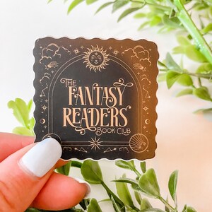 K&ouml;nnte beinhalten: Ein schwarzer und goldener Aufkleber mit einem fantasievollen Design mit Sonne, Mond und Sternen. Der Text "The Fantasy Readers Book Club" ist in einer dekorativen Schrift geschrieben.