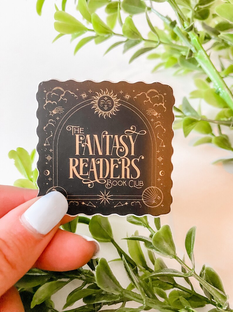 K&ouml;nnte beinhalten: Ein schwarzer und goldener Aufkleber mit einem fantasievollen Design mit Sonne, Mond und Sternen. Der Text "The Fantasy Readers Book Club" ist in einer dekorativen Schrift geschrieben.