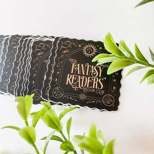 K&ouml;nnte beinhalten: Ein Stapel schwarzer und goldener Aufkleber mit einem fantasievollen Design. Die Aufkleber zeigen den Text "The Fantasy Readers Book Club" mit einem Halbmond, Sternen und Wolken.