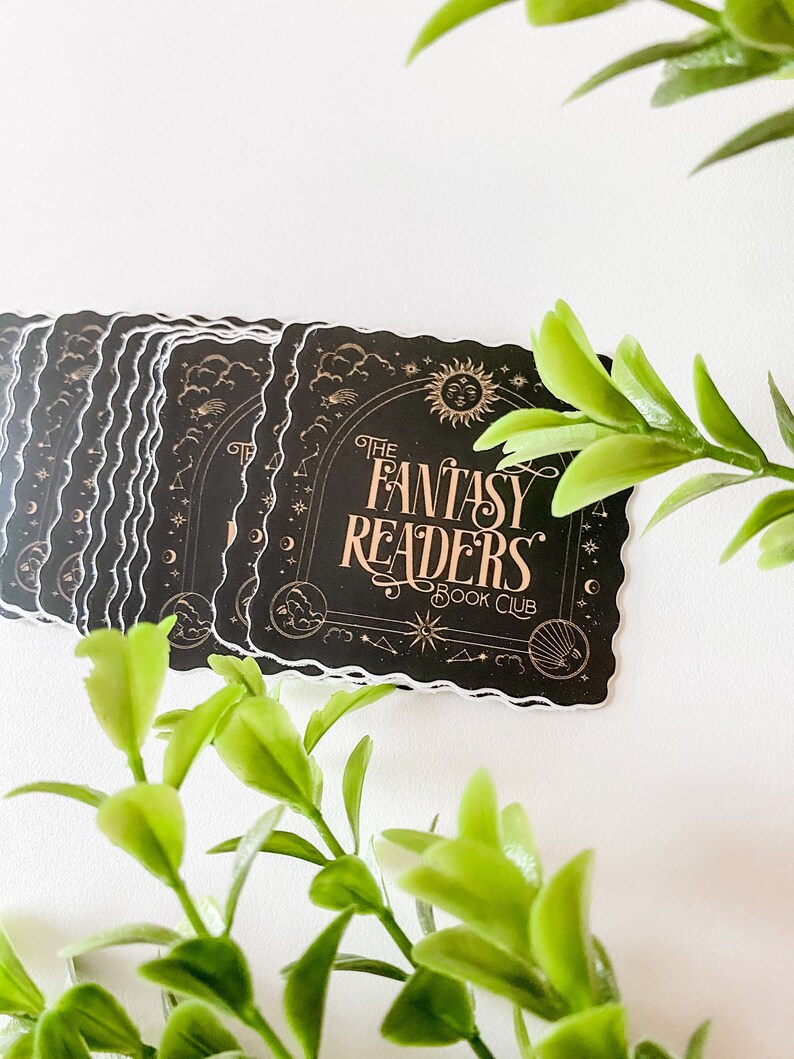 K&ouml;nnte beinhalten: Ein Stapel schwarzer und goldener Aufkleber mit einem fantasievollen Design. Die Aufkleber zeigen den Text "The Fantasy Readers Book Club" mit einem Halbmond, Sternen und Wolken.