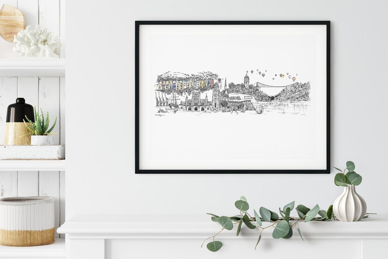 May include: Black and white line drawing of a city skyline with hot air balloons in the sky. The drawing is framed in a black frame and hanging on a white wall.