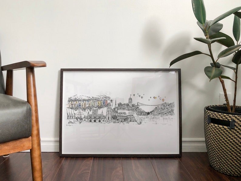 May include: A black and white line drawing of a city skyline with hot air balloons in the sky. The drawing is framed in a black frame and is hanging on a white wall.
