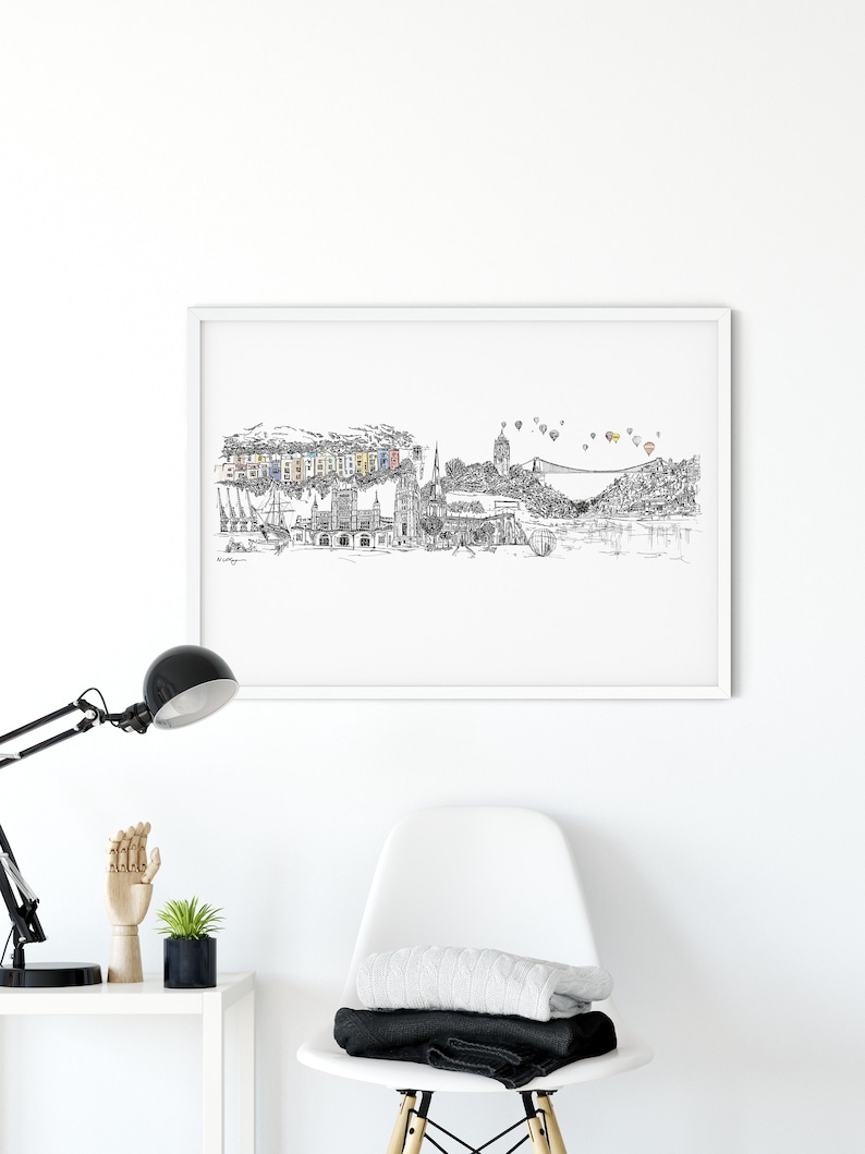 May include: Black and white line drawing of a city skyline with a bridge, buildings, and hot air balloons in the sky. The drawing is framed in a white frame and hanging on a white wall.
