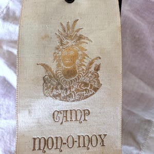 May include: Cream-colored tag with a gold illustration of a person wearing a headdress. The text "CAMP mon-o-moy" is printed in brown, along with a decorative line. A gold cord is attached at the top.
