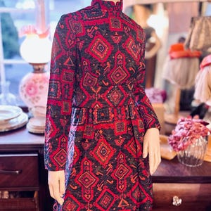 May include: A vintage dress with a high collar and long sleeves, featuring a repeating geometric pattern in red, black, and orange. The dress is paired with a matching red hat. The mannequin is displayed in a shop setting.