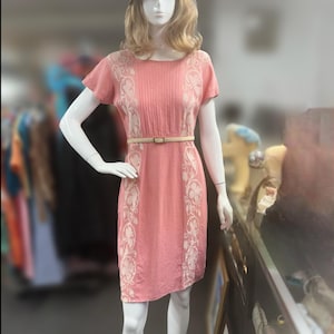 May include: A coral-colored dress with white floral embroidery and a matching belt. The dress has short sleeves and a knee-length hem. A straw hat with a purple band sits atop the mannequin's head.