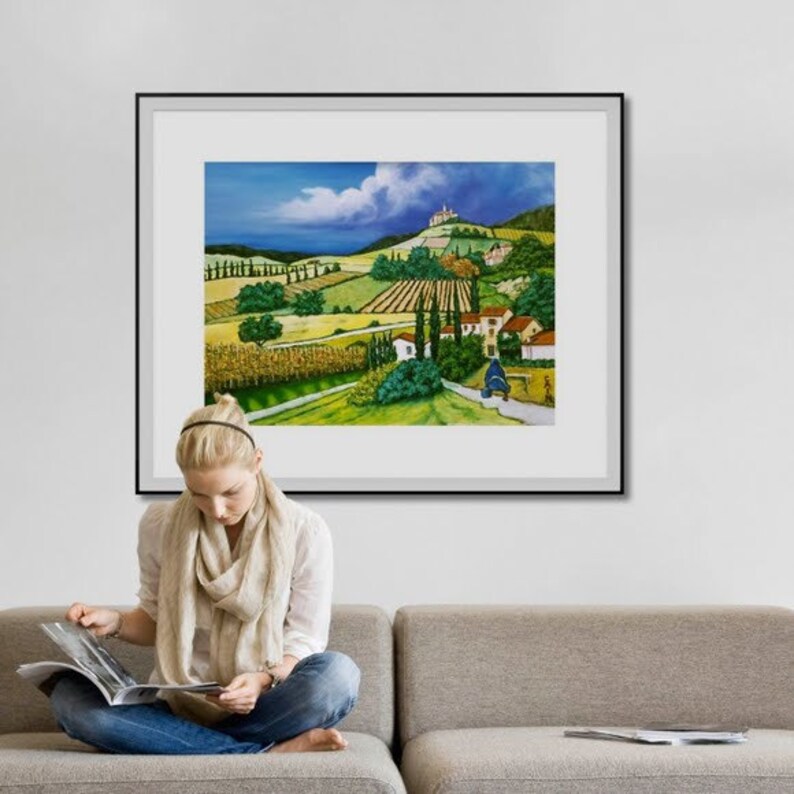 Op de afbeelding: Een ingelijst schilderij van een landelijk landschap met glooiende heuvels, groene velden en een klein dorp in de verte. De lucht is blauw met witte wolken en een vleugje paars in de verte.