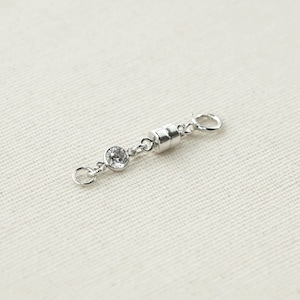 Peut inclure: Fermoir magnétique argenté avec un petit cristal clair rond à une extrémité. Le fermoir a un centre magnétique cylindrique et de petites boucles à chaque extrémité pour la fixation aux bijoux. Fond en tissu beige clair.