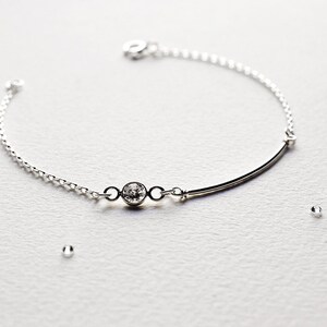 May include: A delicate silver bracelet with a single clear gemstone and a thin silver bar. The bracelet is on a white surface.