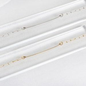May include: Two delicate chain necklaces with a simple bar pendant. The top necklace is silver and the bottom necklace is gold.