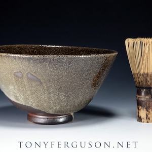 May include: A speckled ceramic tea bowl with a dark rim and base, next to a bamboo whisk. The bowl has a wide, asymmetrical shape with a textured, speckled surface. The whisk is made of thin bamboo tines bound together.