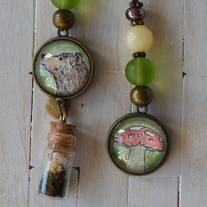 May include: Two handmade beaded bookmarks. One features a bear illustration, a small glass bottle with dried plants, and green, yellow, and brown beads. The other has a mushroom illustration and similar beads. Both have a brown cord.