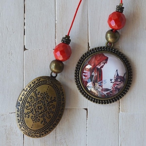 May include: Two antique bronze-toned pendants with red beaded accents. One pendant is oval with an ornate design, the other is round with a picture of Little Red Riding Hood and the wolf. The pendants hang from red cords.