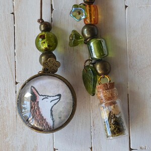 May include: Two handmade charm pendants. One features a fox illustration in a round, bronze-toned frame. The other has a small glass bottle filled with dried herbs, and a decorative bead strand with green and orange beads.