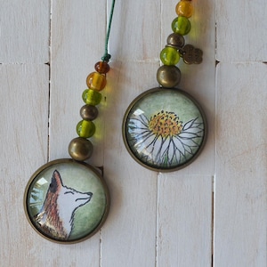 May include: Two round pendants with bronze-colored frames and green cords. One pendant features a fox illustration, the other a white daisy. Each pendant has a string of colorful beads. The background is a white, wooden surface.