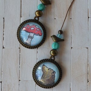 May include: Two bronze-toned pendants with nature-themed illustrations. One pendant features a red mushroom with white spots, and the other depicts a bear looking up. Each pendant is adorned with turquoise beads and a small bird charm.