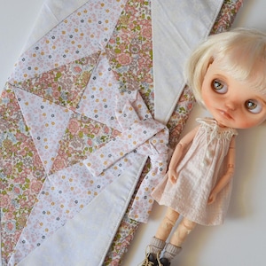 Pode incluir: Uma boneca loira com um vestido rosa está ao lado de uma colcha com estampa floral. A colcha apresenta um design de patchwork com tecido floral branco e rosa. Uma gravata borboleta combinando também está presente. A boneca usa sapatos marrons e meias cinzentas.