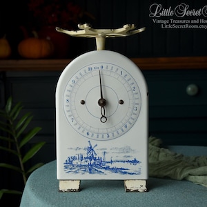 Op de afbeelding: Vintage witte en blauwe keukenweegschaal met een molenlandschap. De weegschaal heeft een ronde wijzerplaat met cijfers en een naald, en meet tot 10 kg. De weegschaal is van metaal en heeft een verweerde uitstraling.