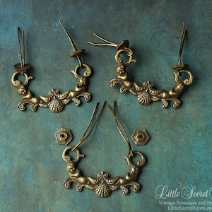 May include: Three brass-toned metal drawer pulls featuring a central shell motif flanked by two cherub figures. Each pull includes two attached metal mounting wires and two separate hexagonal metal hardware pieces for secure installation.